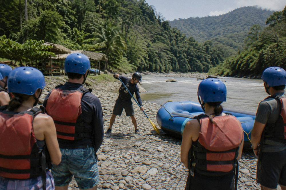 A guide demonstrating paddle strokes to a group wearing helmets and life jackets beside an inflated raft