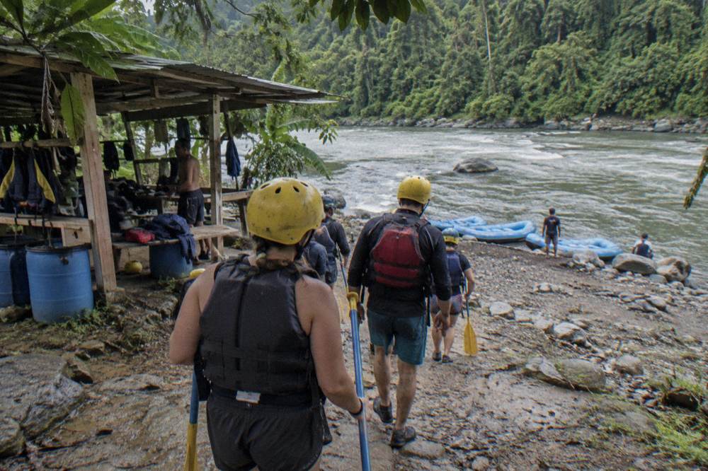 A group of rafters in helmets and life jackets carrying paddles down a rocky riverbank toward a raft