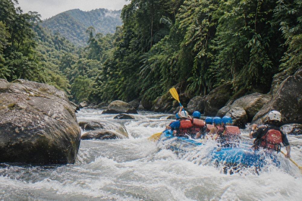 A paddling crew in helmets and life jackets riding a rapid between boulders