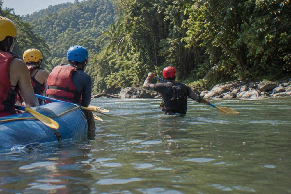 A guide in a red helmet demonstrating paddle strokes to a seated raft crew on a calm river pool