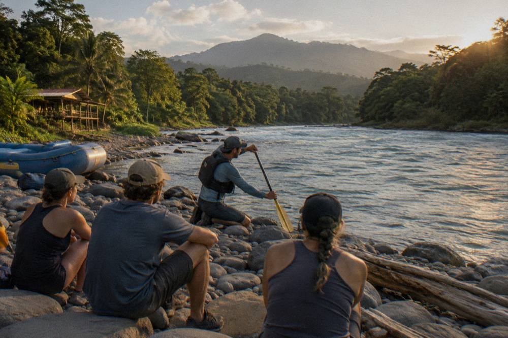 A guide demonstrating paddle strokes to guests on a gravel bank beside turquoise river water