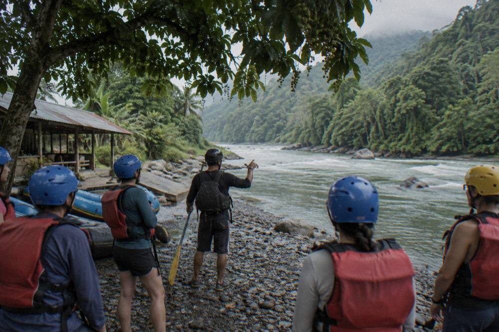 A rafting guide giving a paddle briefing to guests wearing helmets and life jackets on a riverbank