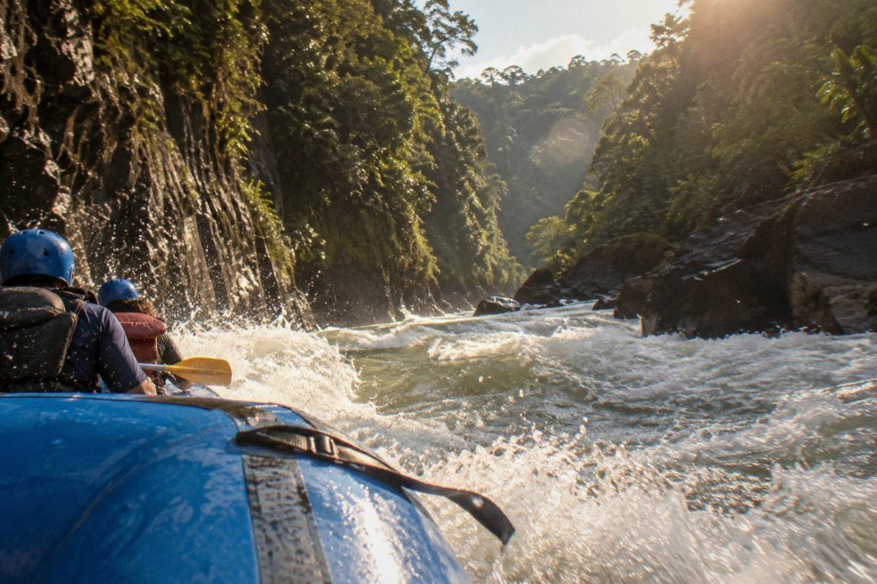 A raft cutting through a green jungle canyon rapids with spray rising off the water