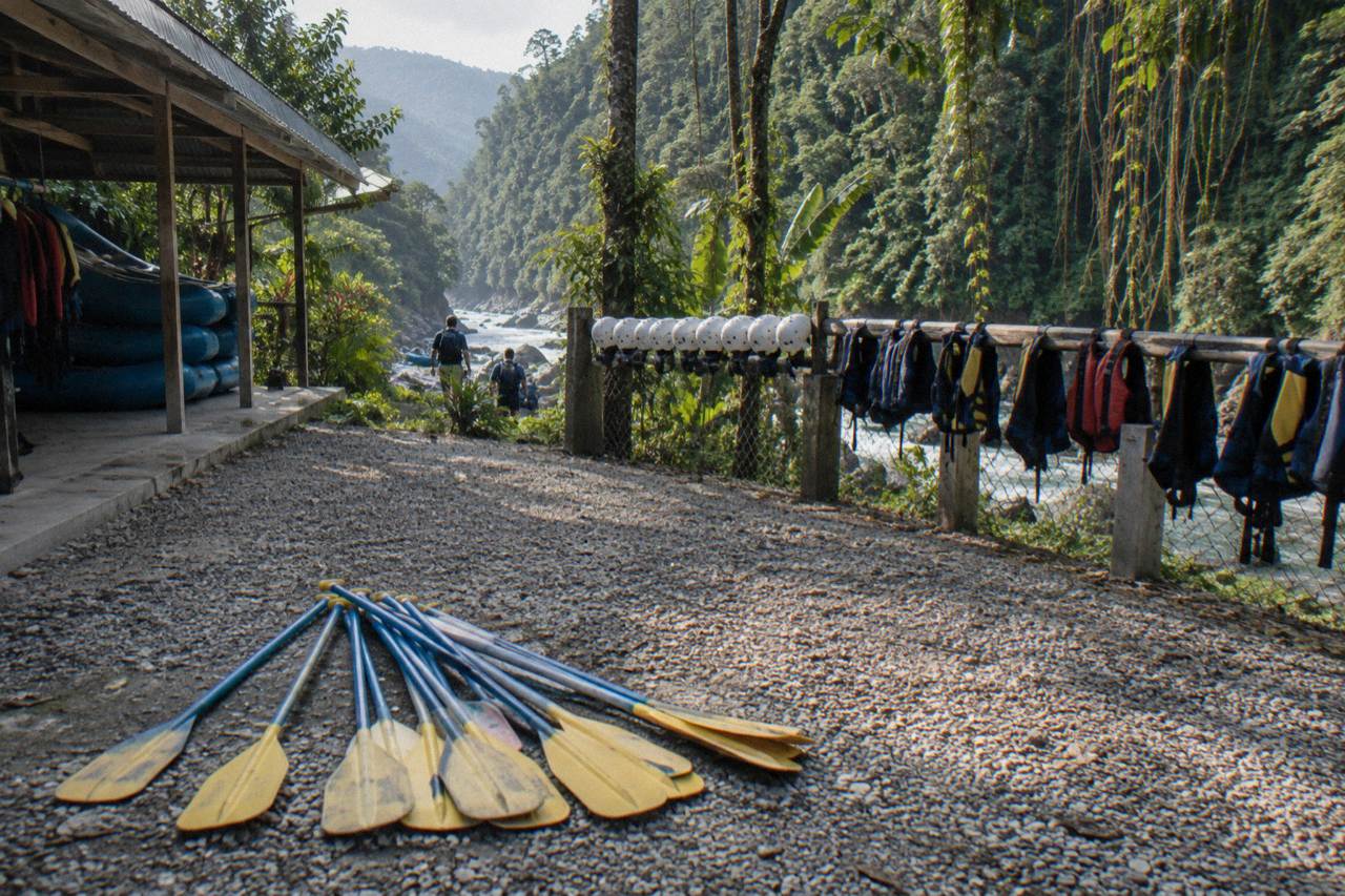 Rafting paddles, helmets and life jackets laid out on a riverbank beside a rainforest gorge