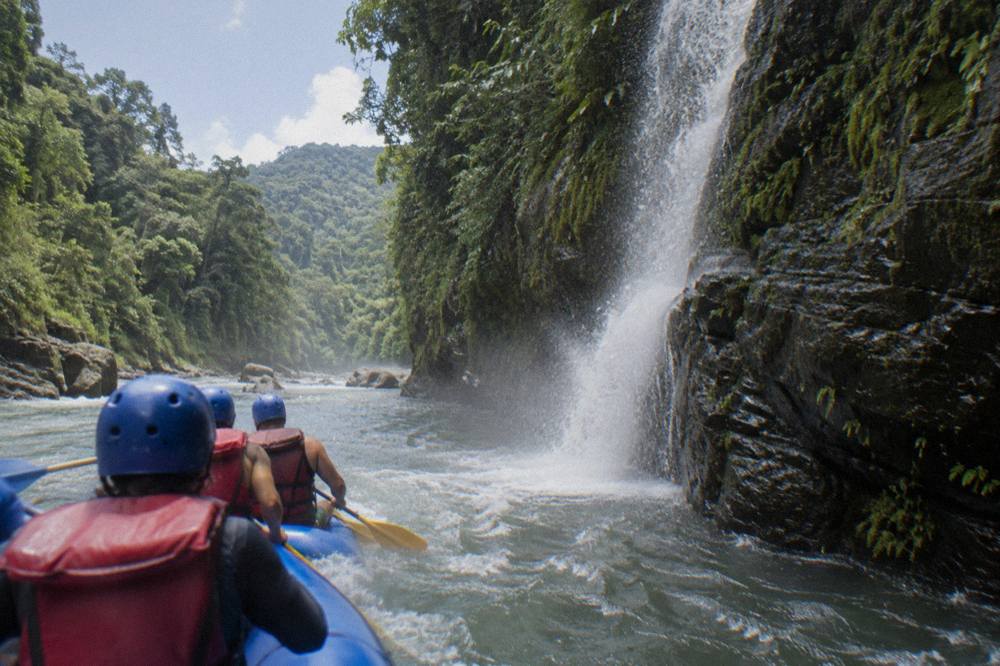 A raft passing a waterfall pouring off a mossy canyon wall into the river