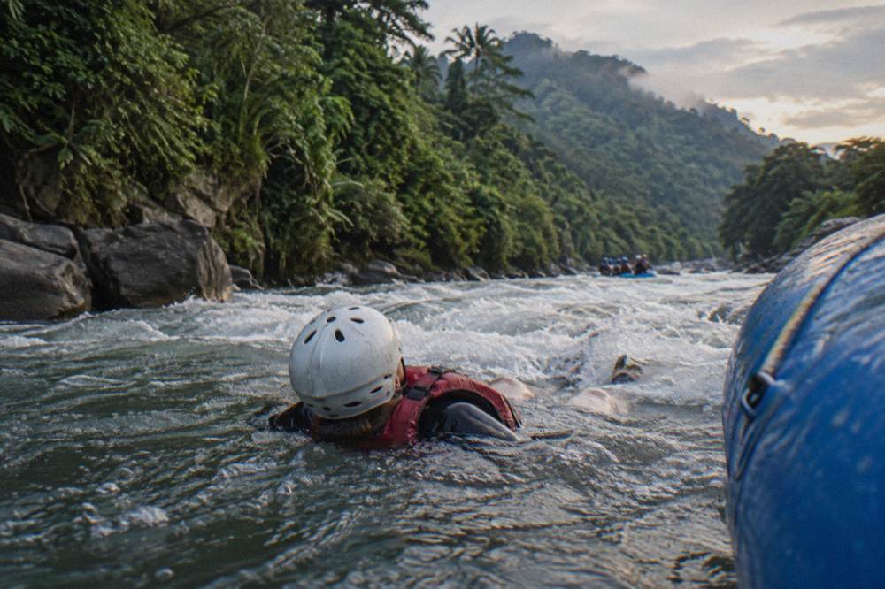A rafter in helmet and life vest floating on their back in a foamy rapid beside the raft