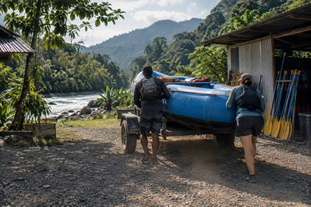 Rafting guides loading a raft and paddles at a gravel river bank in the morning