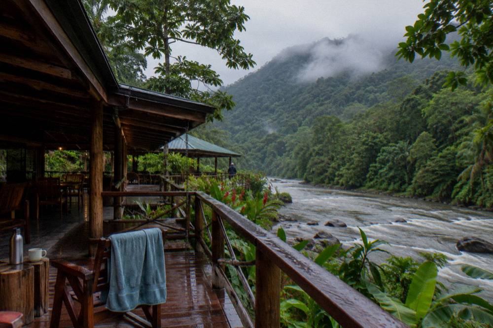 A riverside wooden lodge under rainforest canopy with low cloud on the ridge