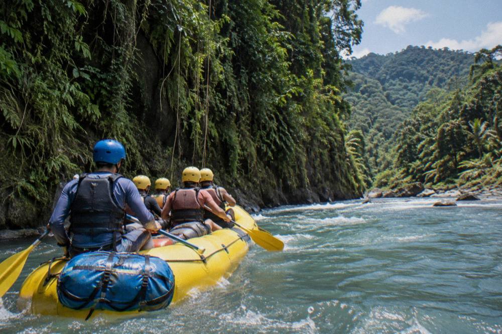 Yellow raft loaded with paddlers passing a sheer rainforest wall above the river