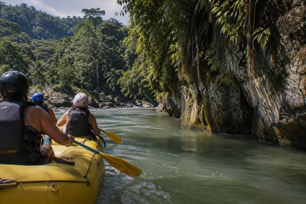 A yellow raft with paddlers passing a rock wall draped in vines on a tropical river