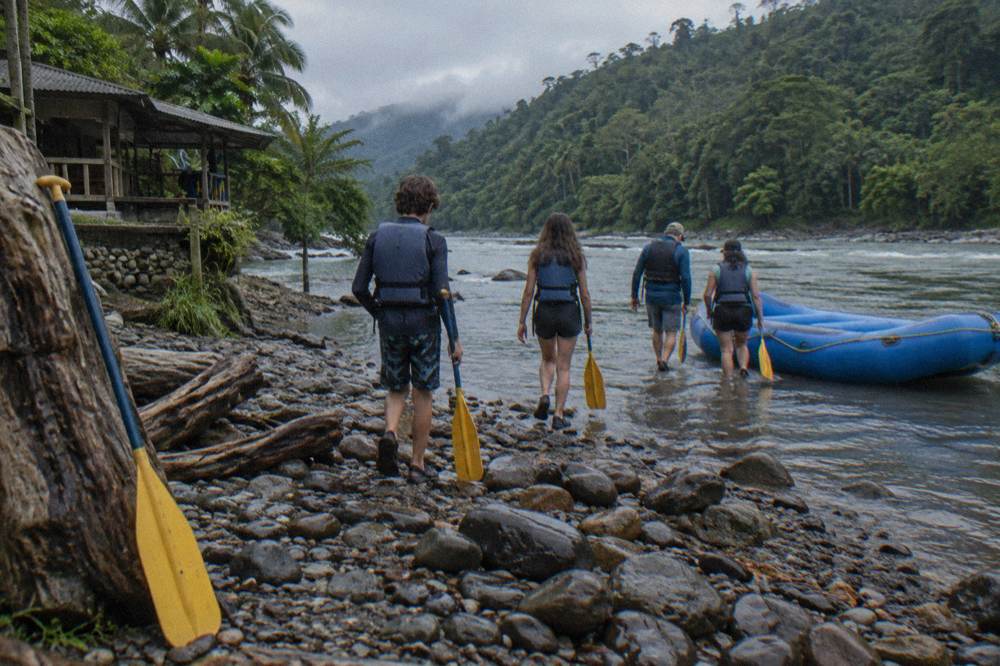 Family with teenagers carrying paddles toward a raft on a gravel riverbank