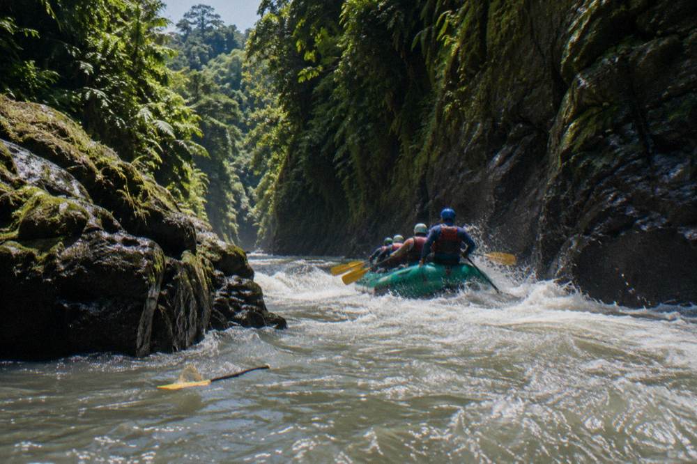 A green raft carrying paddlers through a narrow jungle canyon with spray over the bow