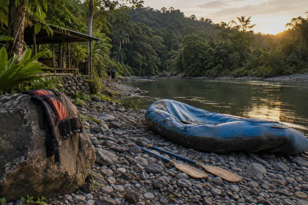 Empty raft and paddles on a rocky riverbank beside calm green water in the late afternoon