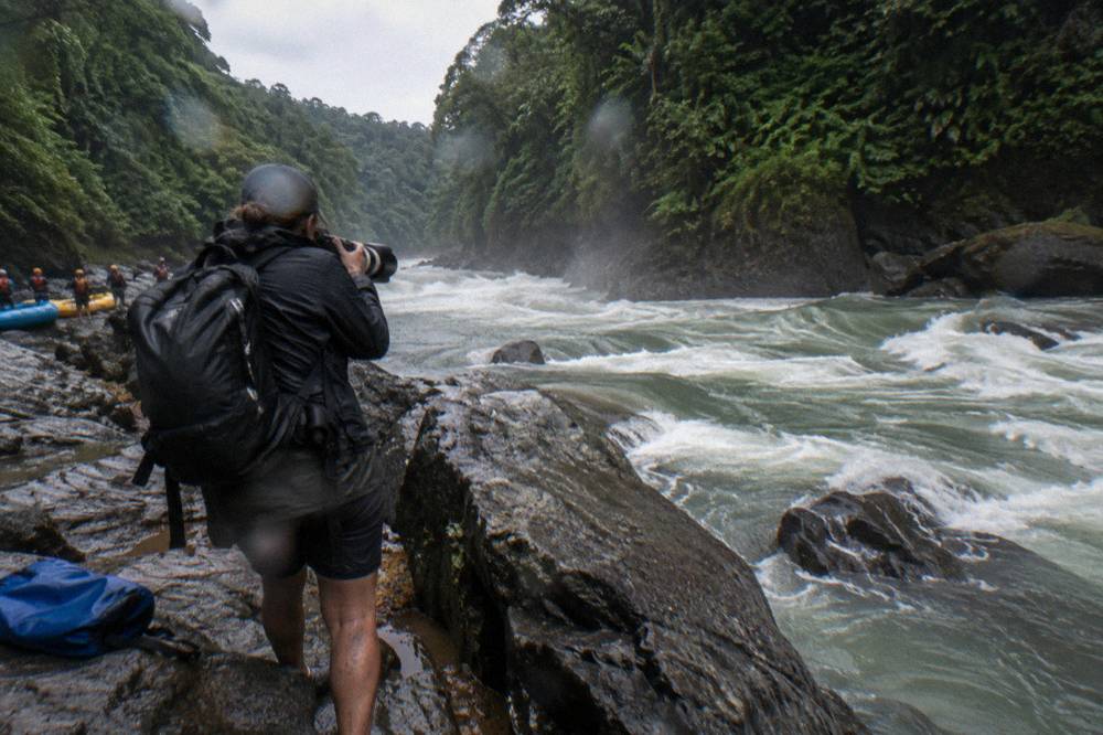 A photographer with a long lens standing on a wet rock beside a rushing rapid