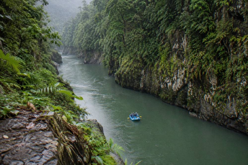 A narrow rainforest gorge with tall green cliffs on both sides and a raft floating on calm water between rapid sections