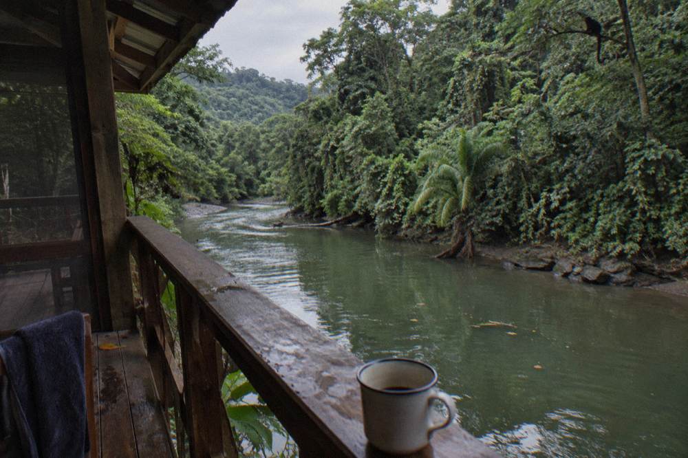 A wooden riverside lodge balcony overlooking a calm green river pool with rainforest on both banks
