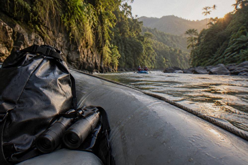 A dry bag and binoculars resting on a raft tube with rainforest and cliffs in the background