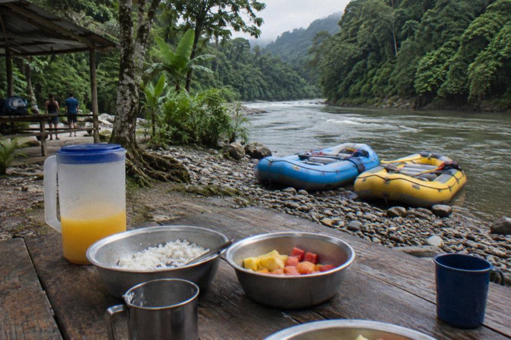 A riverside lunch spread on a wooden table beside rafts pulled up on a pebble bank