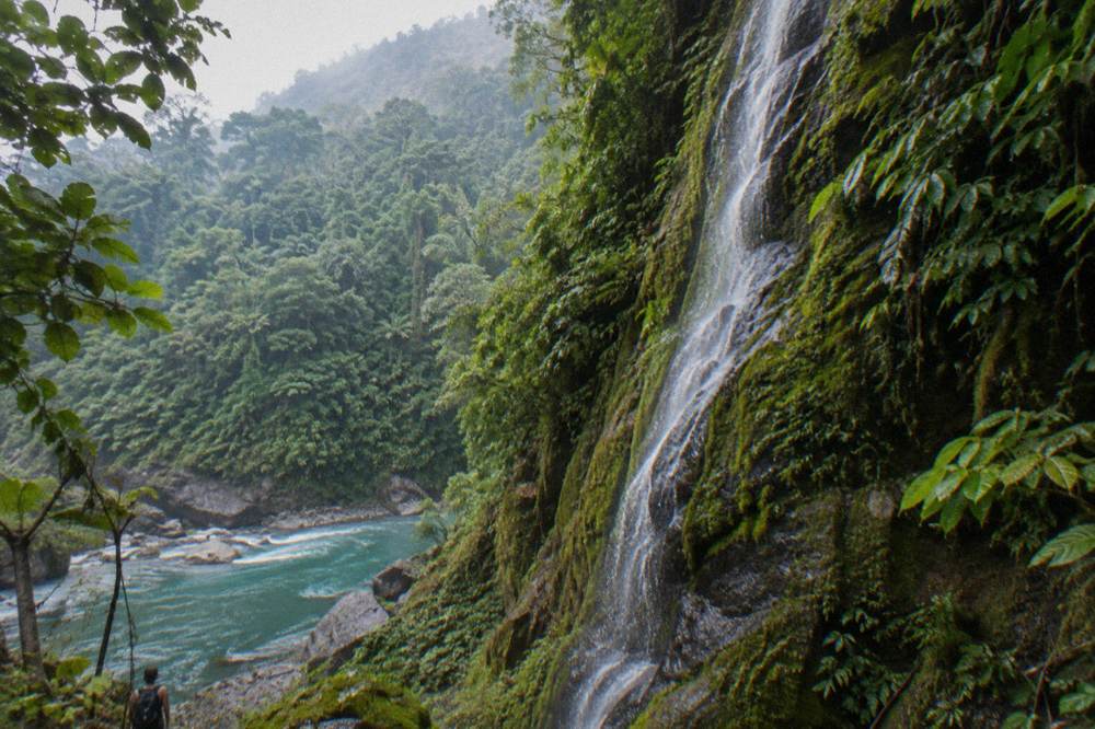 A steep green canyon wall with a thin waterfall falling toward a river far below