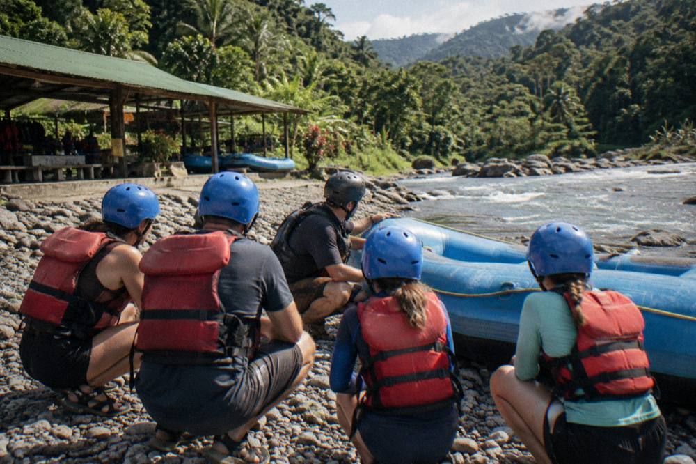 A family in helmets and life jackets listening to a guide beside an inflated raft