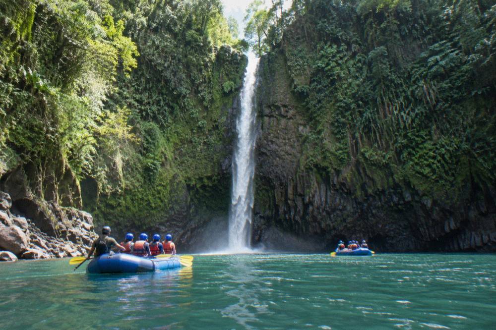 A tall thin waterfall spilling from a mossy cliff directly into a turquoise river between raft trips