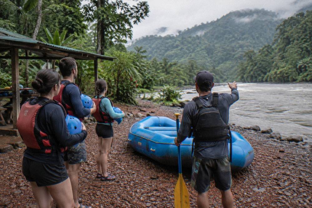 A rafting guide giving a paddle safety talk to guests holding helmets on a gravel riverbank