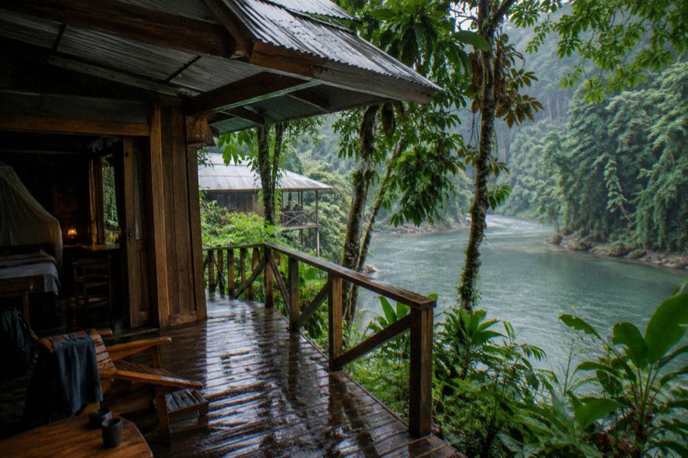A wooden riverside lodge cabin with a metal roof facing the water through tall trees