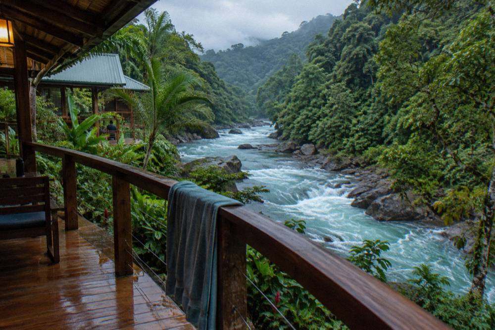 A wooden riverside lodge balcony above turquoise whitewater between dense jungle hills