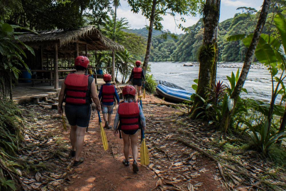 A family in helmets and life jackets carrying paddles along a riverside path
