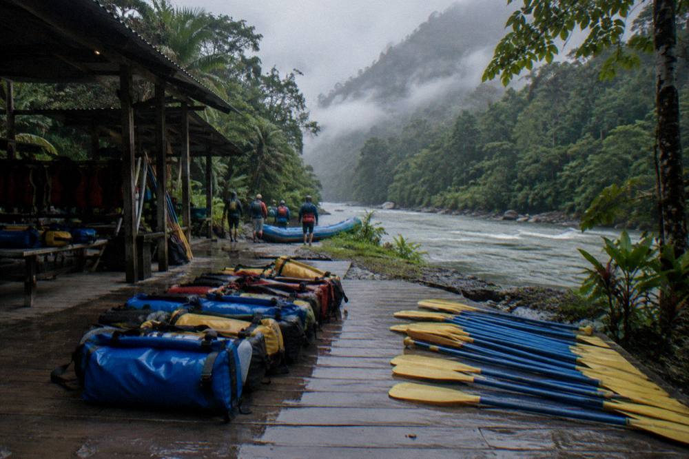 Dry bags and paddles laid out on a riverside platform beside a rainforest trail