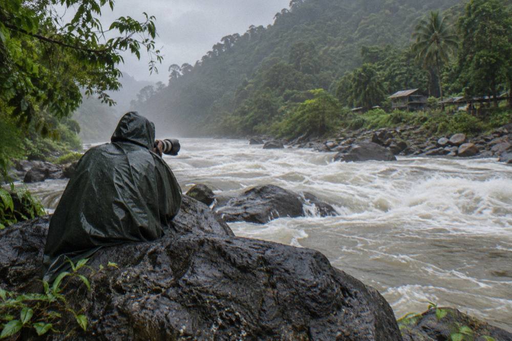 A photographer with a telephoto lens standing on a wet boulder beside a whitewater rapid