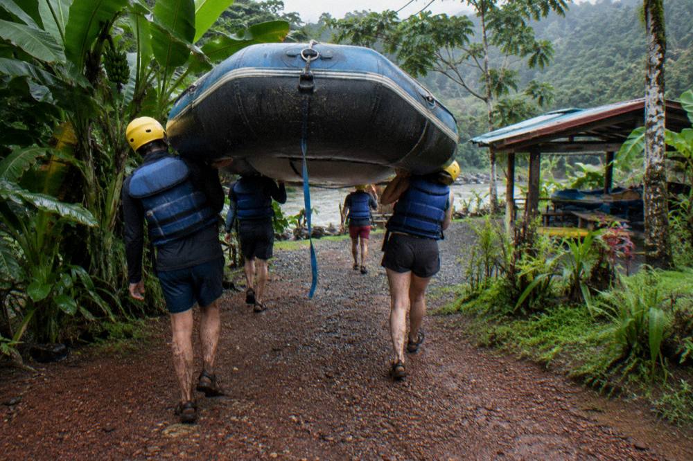 A rafting group in helmets and life jackets carrying a raft toward the river through trees
