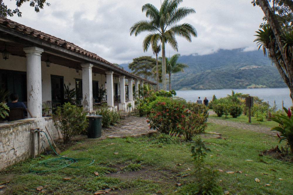 Whitewashed hacienda-style hotel with a columned veranda across a palm garden and a calm lake